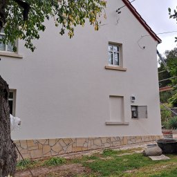 IZO-PLUS PAWEŁ WALSKI - Otynkowana elewacja domu jednorodzinnego z dekoracyjnym kamieniem u podstawy. Widoczne okna, skrzynka elektryczna i fragment drzewa. Świeży tynk w kolorze beżowym.