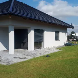 Nowoczesny dom jednorodzinny z ciemną dachówką, jasną elewacją i szarymi roletami zewnętrznymi, otoczony zielonym trawnikiem i ogrodzeniem. Widoczny bus w tle.