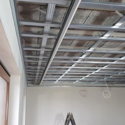 IZO-PLUS PAWEŁ WALSKI - Metalowa konstrukcja sufitu podwieszanego w trakcie montażu, widoczne profile i mocowania. W tle surowy strop i wystające kable instalacji elektrycznej.