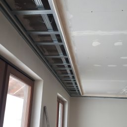 IZO-PLUS PAWEŁ WALSKI - Montaż sufitu podwieszanego z płyty karton-gips na metalowej konstrukcji. Widoczne łączenia płyt i fragment okna. Jasne wnętrze w trakcie prac wykończeniowych.