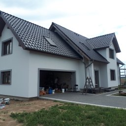 IZO-PLUS PAWEŁ WALSKI - Biały dom jednorodzinny z ciemną dachówką i szarą elewacją po malowaniu. Widoczne farby, drabina i otwarty garaż z osobą w środku. Brukowany podjazd.