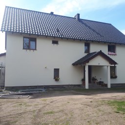 IZO-PLUS PAWEŁ WALSKI - Dwukondygnacyjny dom z ciemną dachówką po malowaniu elewacji, z rusztowaniem i drabinami w pobliżu. Widoczne kwiaty w oknach i zadaszone wejście.