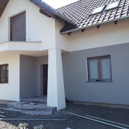 IZO-PLUS PAWEŁ WALSKI - Dwukolorowa elewacja domu jednorodzinnego z widocznymi oknami zabezpieczonymi folią malarską i rusztowaniem w trakcie prac wykończeniowych.