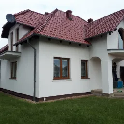 Elewacja domu jednorodzinnego z czerwoną dachówką po malowaniu, widoczna antena satelitarna i pracownik przy kolumnach wejściowych. Zieleń trawnika i częściowo widoczny bus.