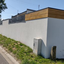 IZO-PLUS PAWEŁ WALSKI - Nowoczesne ogrodzenie z białego tynku z drewnianymi i metalowymi elementami, widok z ulicy, skrzynka elektryczna przy ścianie.