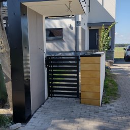IZO-PLUS PAWEŁ WALSKI - Nowoczesne ogrodzenie posesji z czarną, poziomą bramą i słupkami wykończonymi drewnem oraz elementami z szarego kamienia na tle domu z numerem 15B.