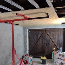 IZO-PLUS PAWEŁ WALSKI - Montaż płyty OSB na suficie garażu za pomocą podnośnika. Widoczne metalowe profile sufitowe i nieotynkowane ściany. W tle brązowa brama garażowa.