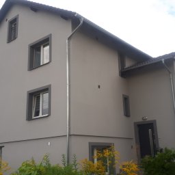 IZO-PLUS PAWEŁ WALSKI - Odnowiona elewacja dwupiętrowego domu w szarościach z ciemnymi obramowaniami okien i dachu, z widocznymi rynnami i zielenią w ogrodzie.