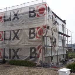 IZO-PLUS PAWEŁ WALSKI - Dom w trakcie ocieplania, osłonięty siatką z logo Bolix, widoczne rusztowania i fragment ogrodu z samochodem. Prace elewacyjne w toku.