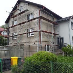 IZO-PLUS PAWEŁ WALSKI - Dom w trakcie renowacji elewacji, otoczony rusztowaniem. Widoczne okna i fragment dachu. W tle zieleń i sąsiedni budynek.