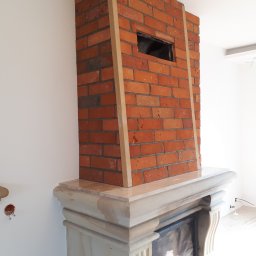 IZO-PLUS PAWEŁ WALSKI - Murowany kominek z czerwonej cegły, obudowany jasnym kamieniem, z widocznym otworem wentylacyjnym i drewnianymi listwami wykończeniowymi, na tle białej ściany.