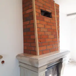 Murowany kominek z czerwonej cegły, obudowany jasnym kamieniem, z widocznym otworem wentylacyjnym i drewnianymi listwami wykończeniowymi, na tle białej ściany.