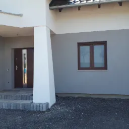 Dwukolorowa elewacja domu z brązowymi oknami i drzwiami wejściowymi. Widoczne schody z kostki brukowej i szara elewacja w dolnej części budynku.