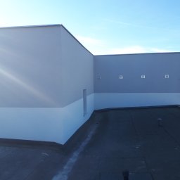 IZO-PLUS PAWEŁ WALSKI - Elewacja budynku po malowaniu z drabinką i małymi okienkami wentylacyjnymi, widok z dachu w słoneczny dzień. Jasnoszara góra i biały dół elewacji.