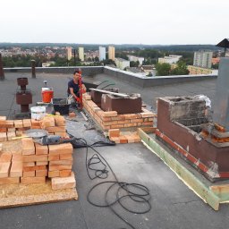 IZO-PLUS PAWEŁ WALSKI - Murarz w trakcie budowy komina na dachu budynku. Widoczne cegły, narzędzia i panorama miasta w tle. Prace murarskie z użyciem poziomicy i zaprawy.