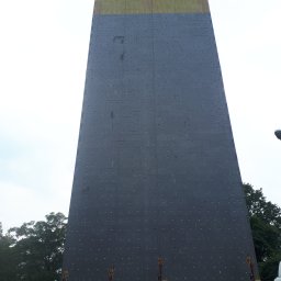 IZO-PLUS PAWEŁ WALSKI - Wysoka ściana budynku w trakcie ocieplania, z widoczną szarą warstwą izolacyjną i górną częścią pokrytą żółtym materiałem izolacyjnym. Prace na wysokości.