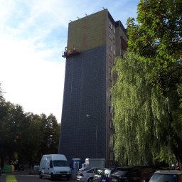 IZO-PLUS PAWEŁ WALSKI - Ocieplanie elewacji budynku wielorodzinnego z użyciem rusztowania wiszącego; widoczne płyty izolacyjne i siatka z otworami. Na dole parkujące samochody i drzewo.