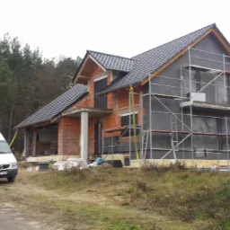 Dom w trakcie ocieplania ścian zewnętrznych, widoczne rusztowania, częściowo ocieplona elewacja, cegła klinkierowa i zaparkowany biały bus.