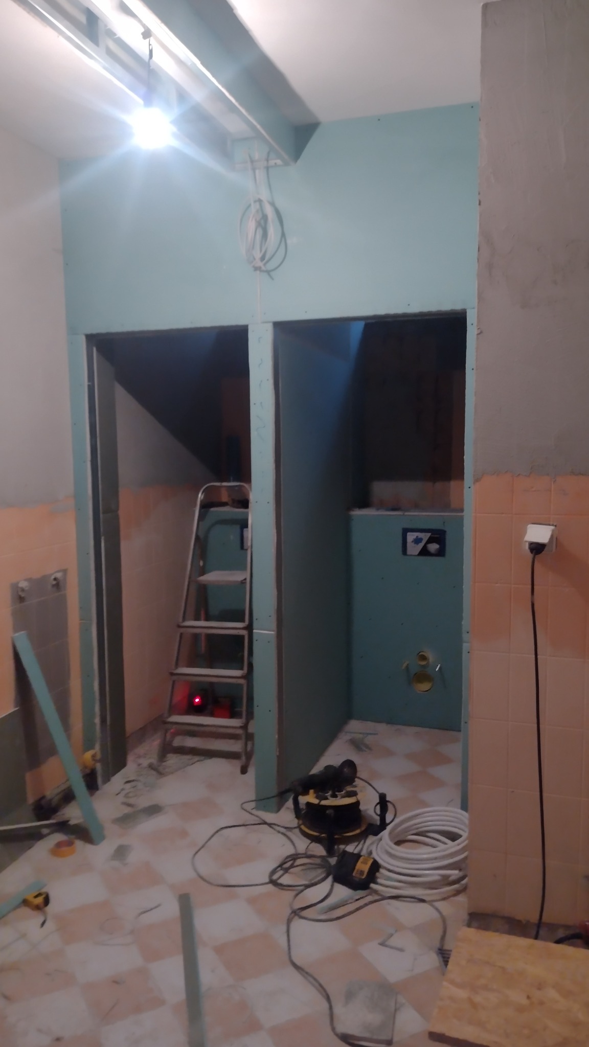 Wnętrze w trakcie remontu: ściany z płyt karton-gips, drabina, narzędzia i przewody na podłodze w kratkę. Widoczne elementy instalacji.