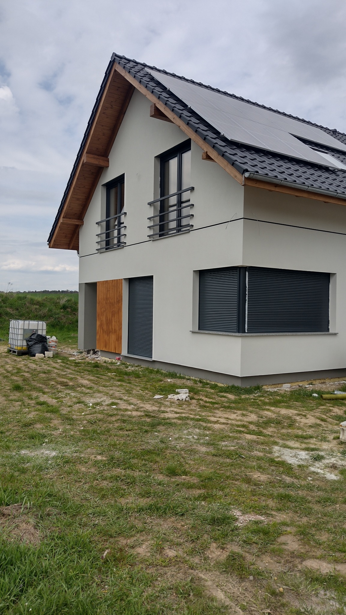 Nowoczesny dom z szarą elewacją, drewnianym wykończeniem i panelami słonecznymi na dachu. Widoczne rolety zewnętrzne i balkon z metalową balustradą.