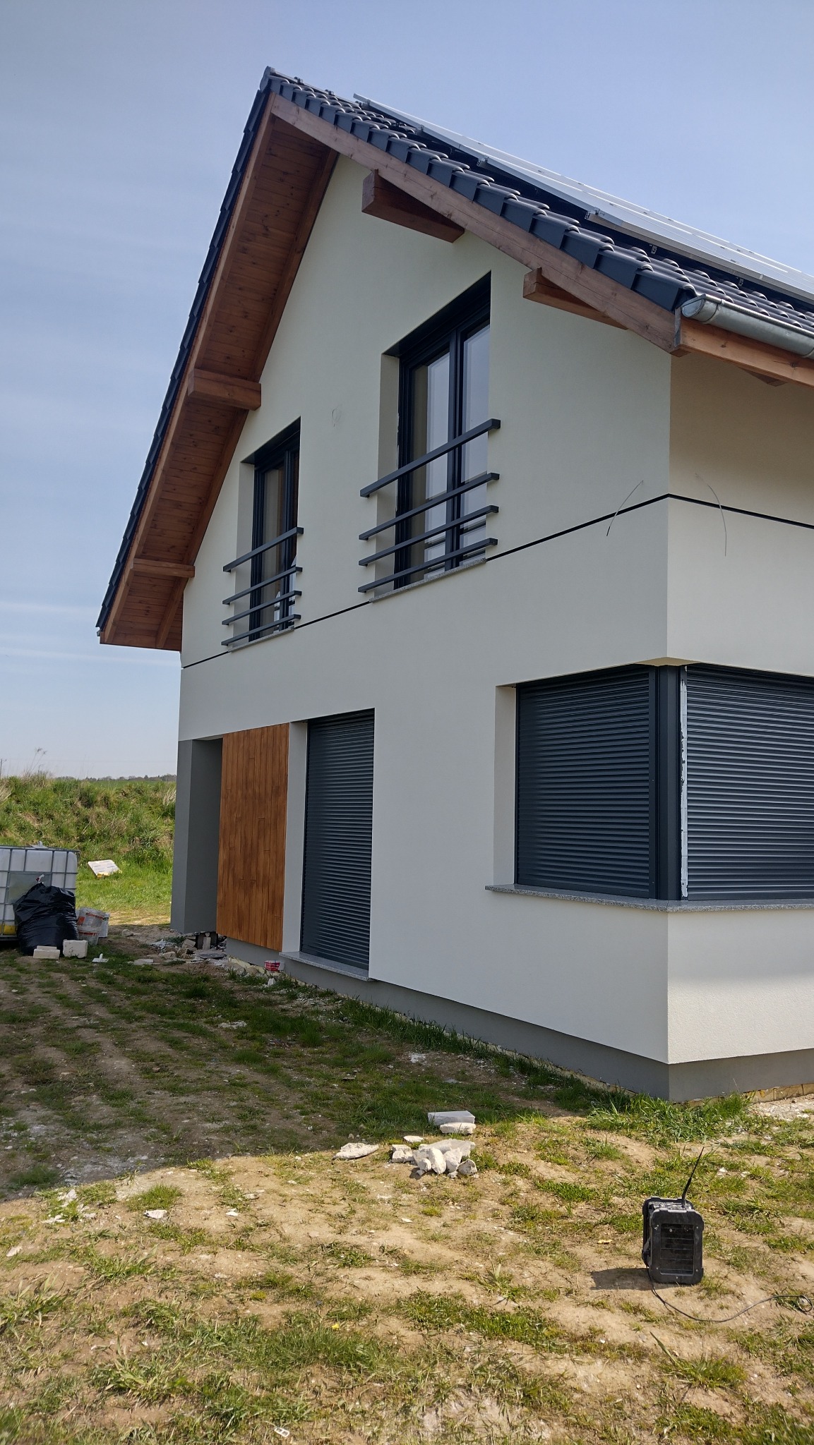 Nowoczesny dom z jasną elewacją, grafitowymi roletami i drewnianymi akcentami. Widoczne wykończenia i detale budynku, w tle zielona trawa i błękitne niebo.