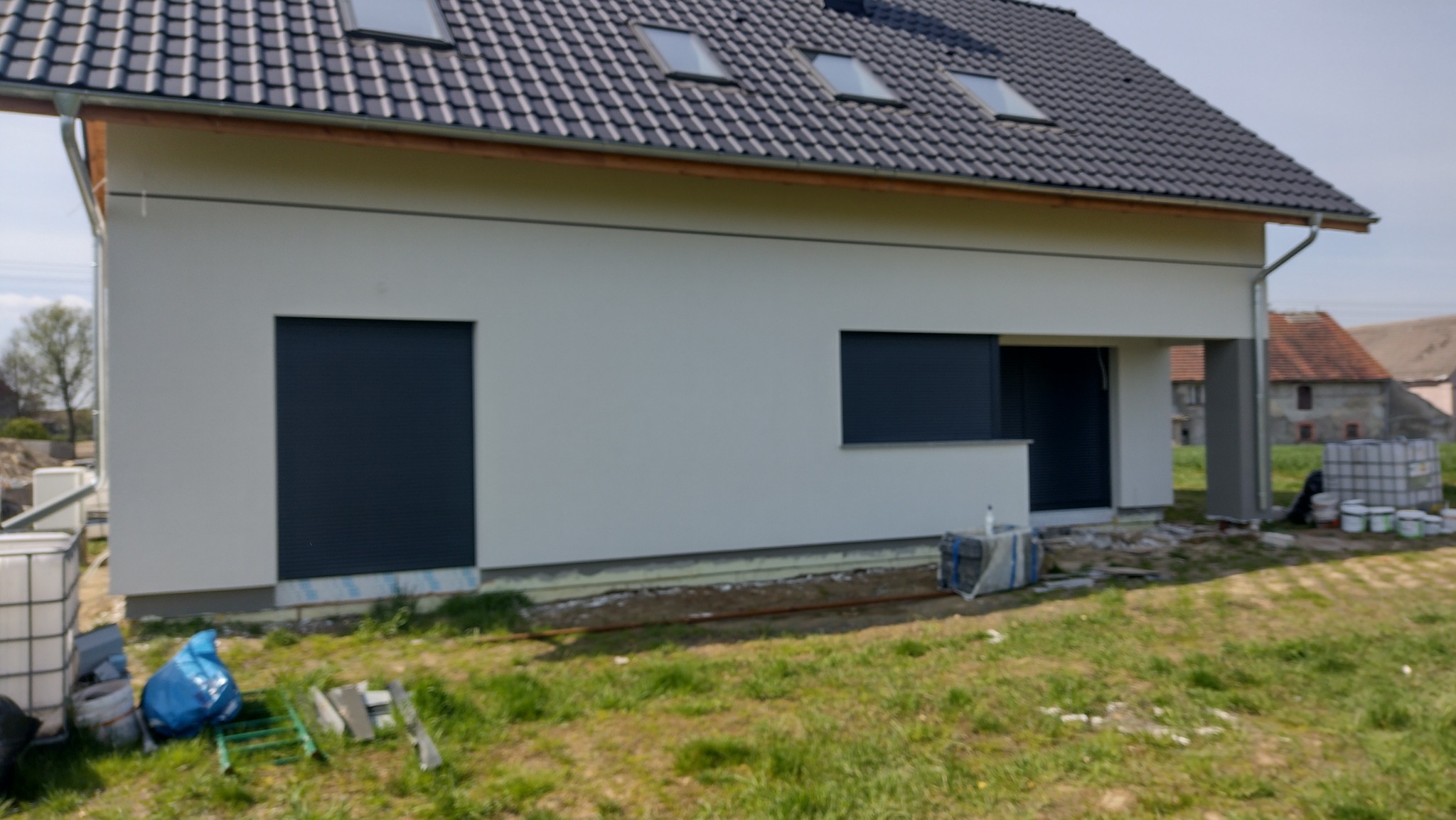 Boczny widok domu z białą elewacją, grafitowym dachem z oknami połaciowymi i ciemnymi roletami. Na trawie widoczne materiały budowlane i puszki po farbach.