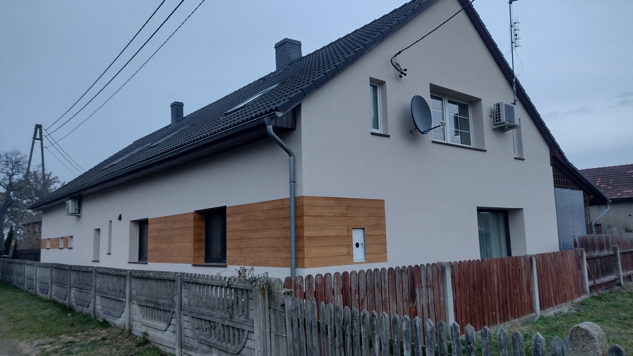 Wykończony dom z ocieploną elewacją, fragmentami wykończonymi drewnopodobną okładziną. Widoczny fragment ogrodzenia i satelita na ścianie.