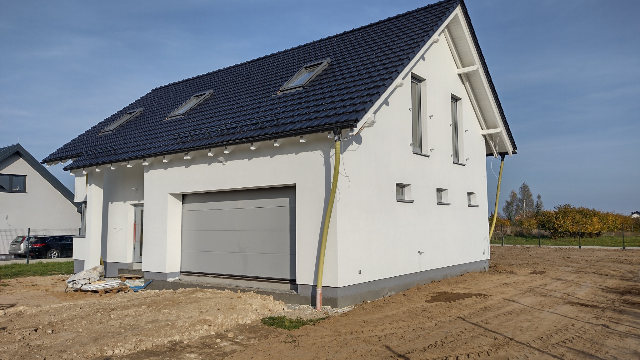 Nowoczesny, biały dom jednorodzinny z grafitowym dachem i szarą bramą garażową. Widoczny fragment elewacji i otoczenie w trakcie prac wykończeniowych.