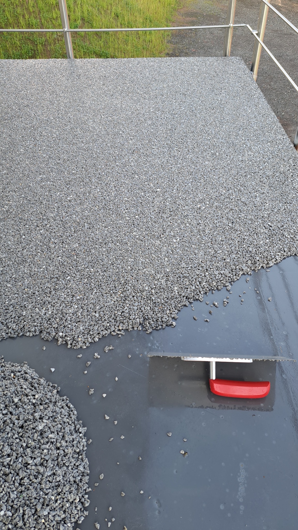 Aplikacja żywicy z kamyczkami na balkonie, widoczna szpachelka z czerwoną rączką. Część powierzchni pokryta, reszta przygotowana. Balustrada w tle.
