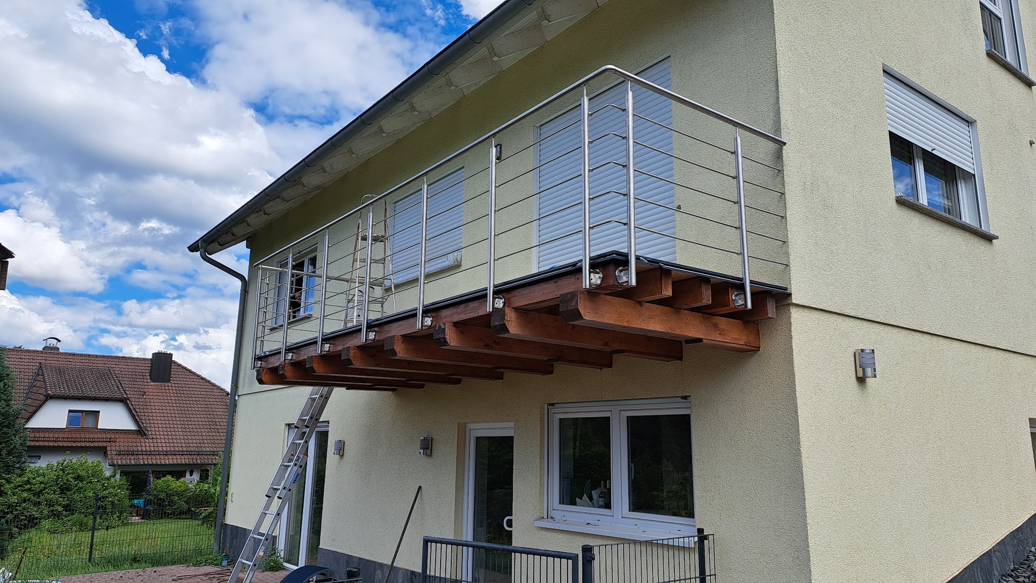 Nowoczesny balkon z drewnianą konstrukcją i metalową balustradą, widok z dołu, drabina oparta o ścianę, dom z elewacją w tle.