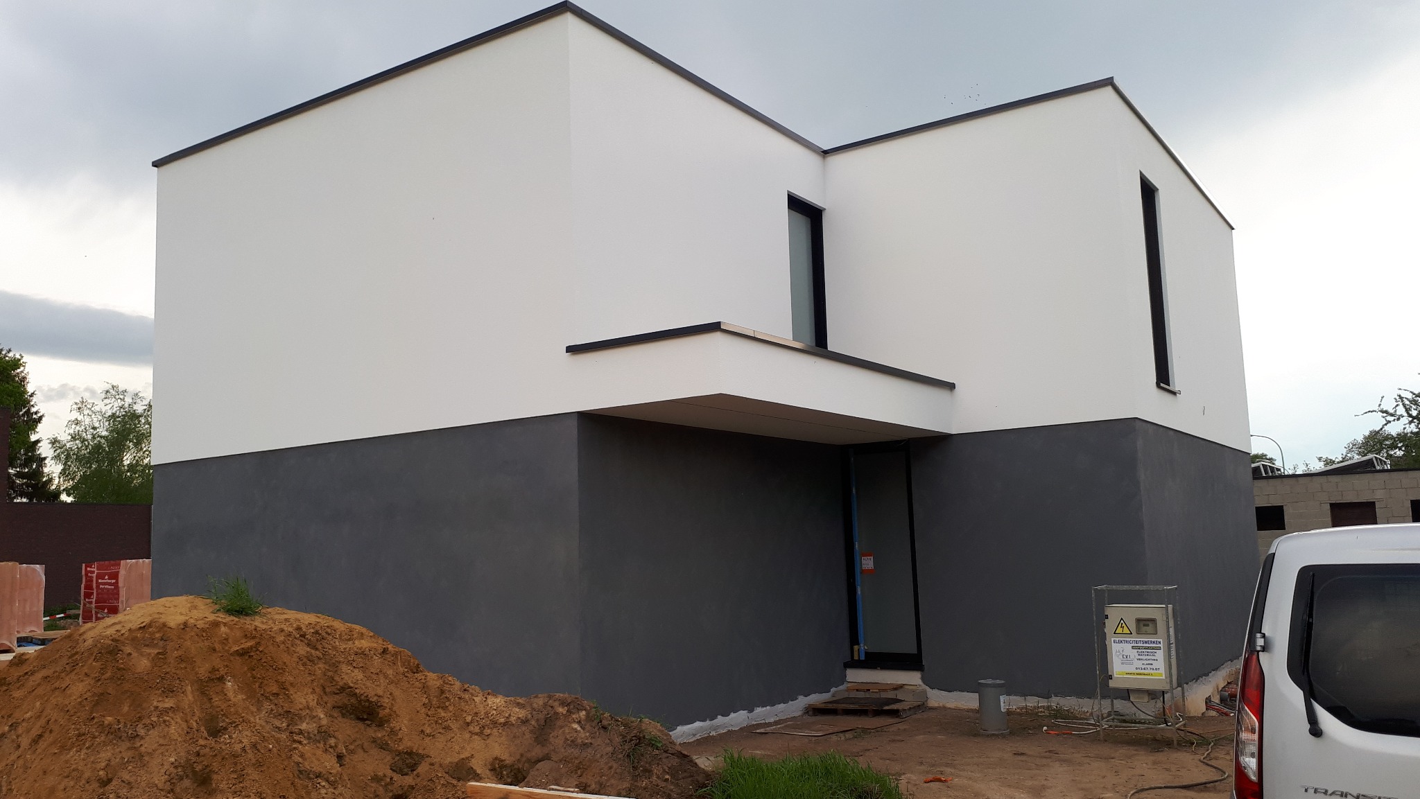 Nowoczesny dom z elewacją w dwóch odcieniach szarości i bieli, z minimalistycznymi oknami. Na pierwszym planie hałda ziemi i fragment samochodu dostawczego.