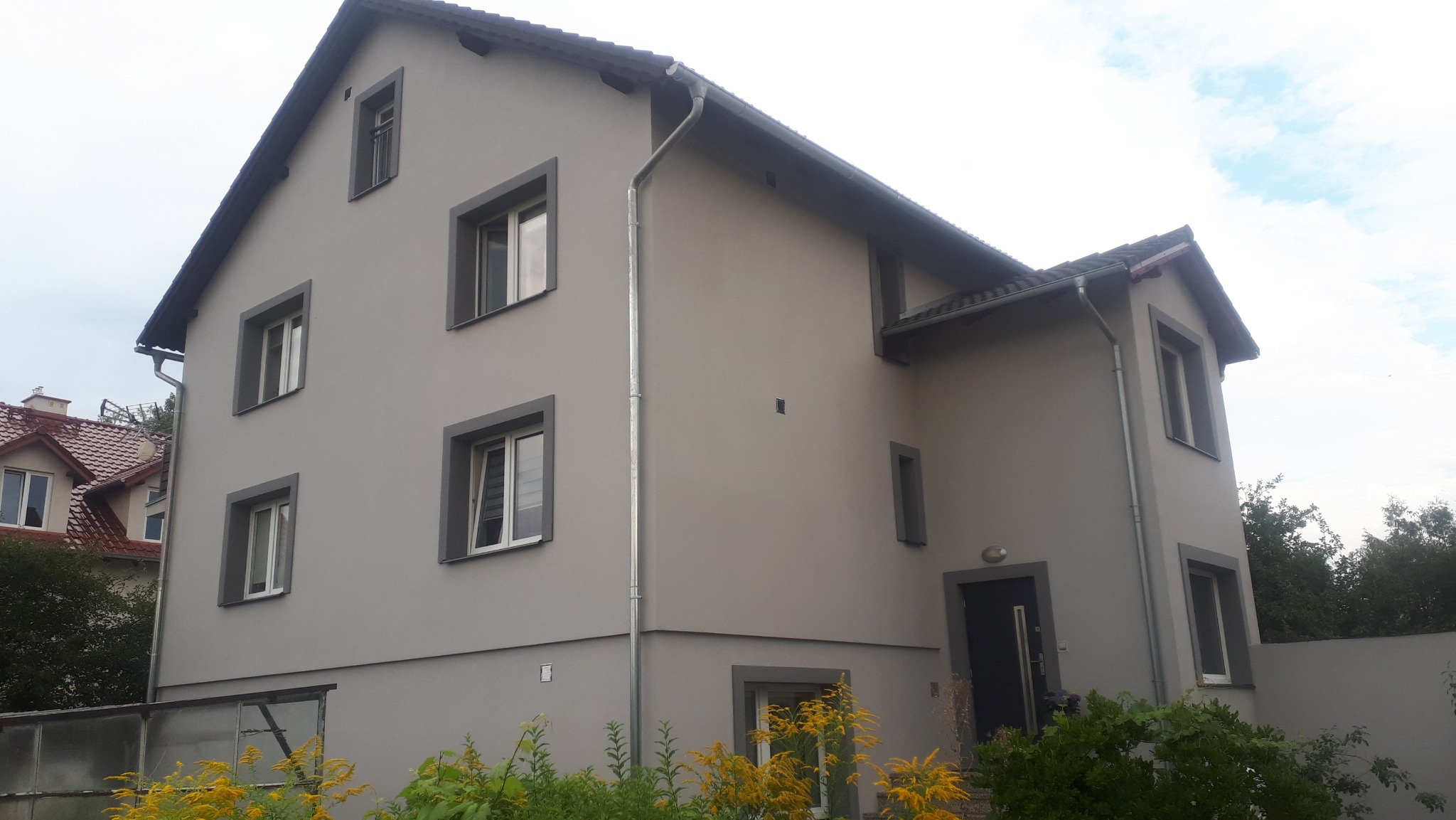 Odnowiona elewacja dwupiętrowego domu w szarościach z ciemnymi obramowaniami okien i dachu, z widocznymi rynnami i zielenią w ogrodzie.