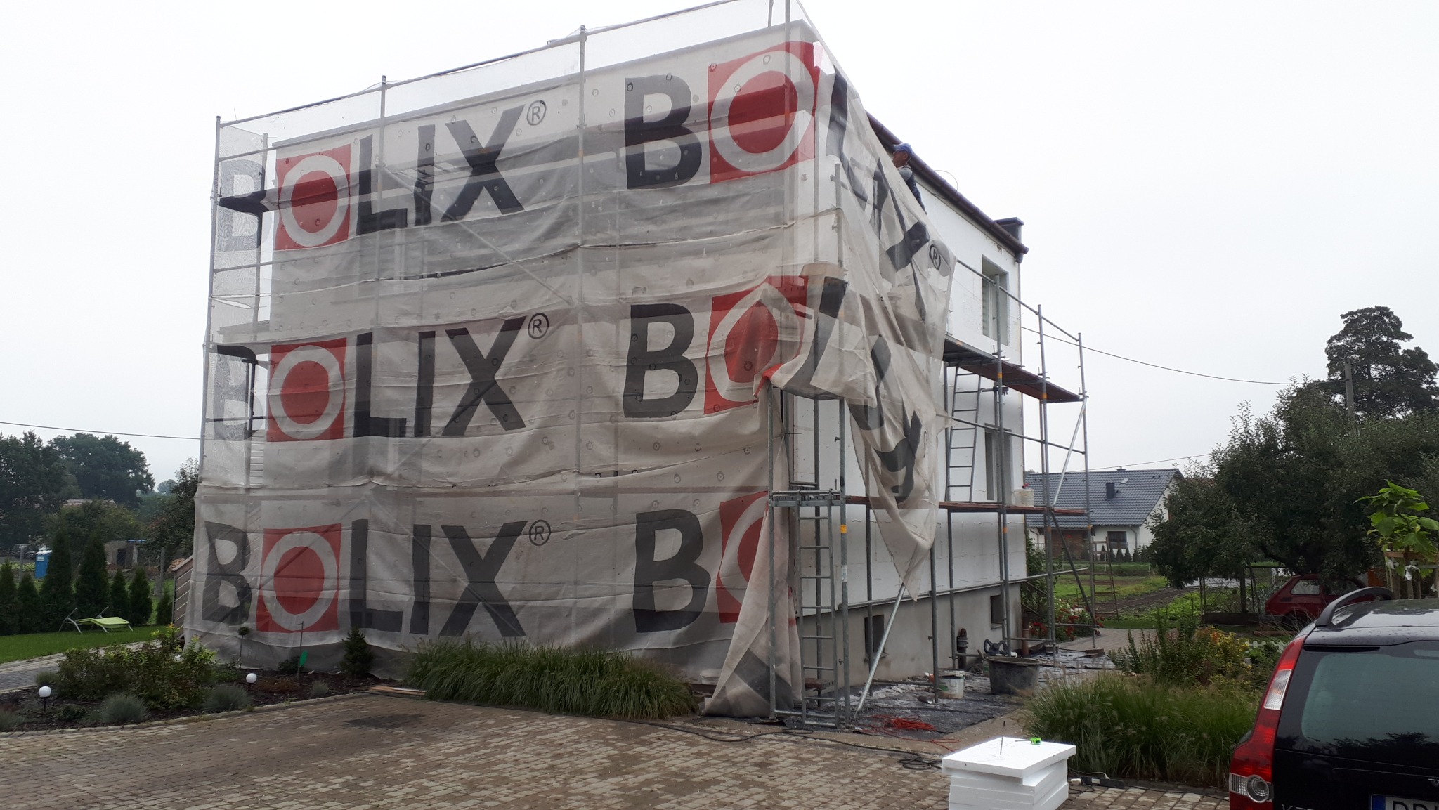 Dom w trakcie ocieplania, osłonięty siatką z logo Bolix, widoczne rusztowania i fragment ogrodu z samochodem. Prace elewacyjne w toku.