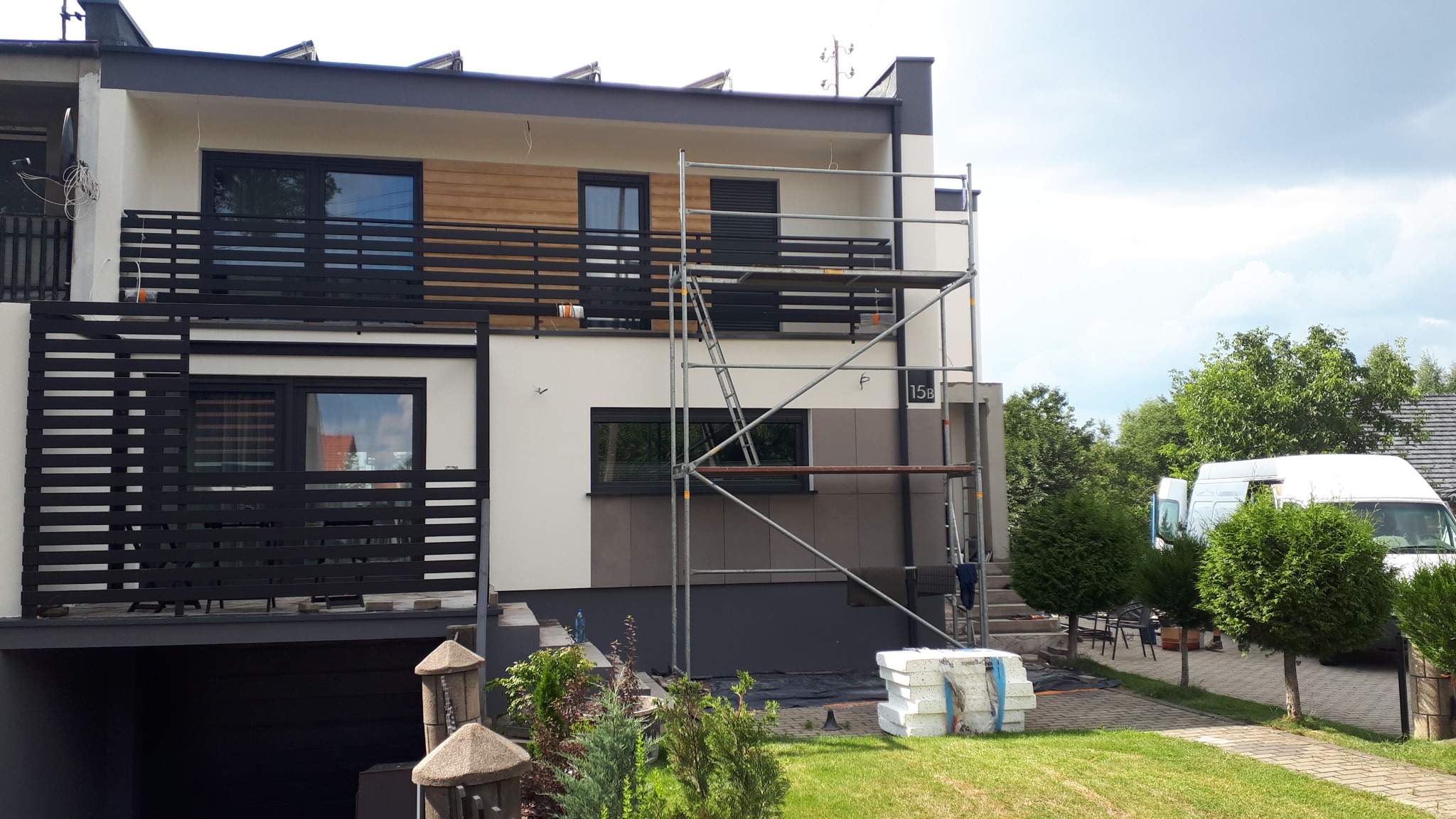 Dwukondygnacyjny dom w trakcie elewacji, widoczne rusztowania, bloki styropianu, częściowo wykończona fasada w odcieniach bieli i szarości, na balkonie drewniana okładzina.