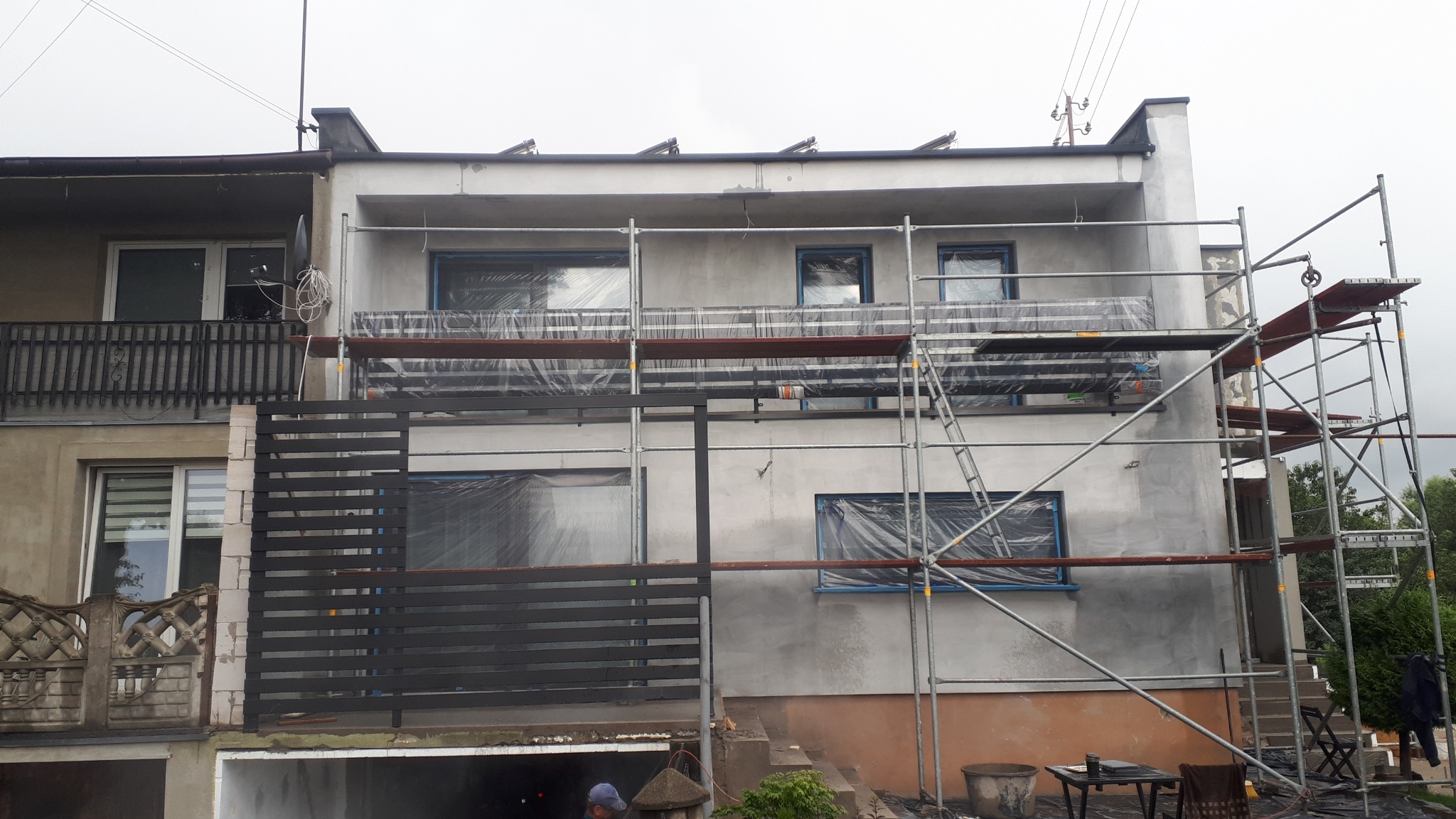 Dom w trakcie ocieplania z rusztowaniem i folią ochronną na oknach. Widoczna świeżo nałożona warstwa izolacyjna, sąsiadujący budynek z balkonem.