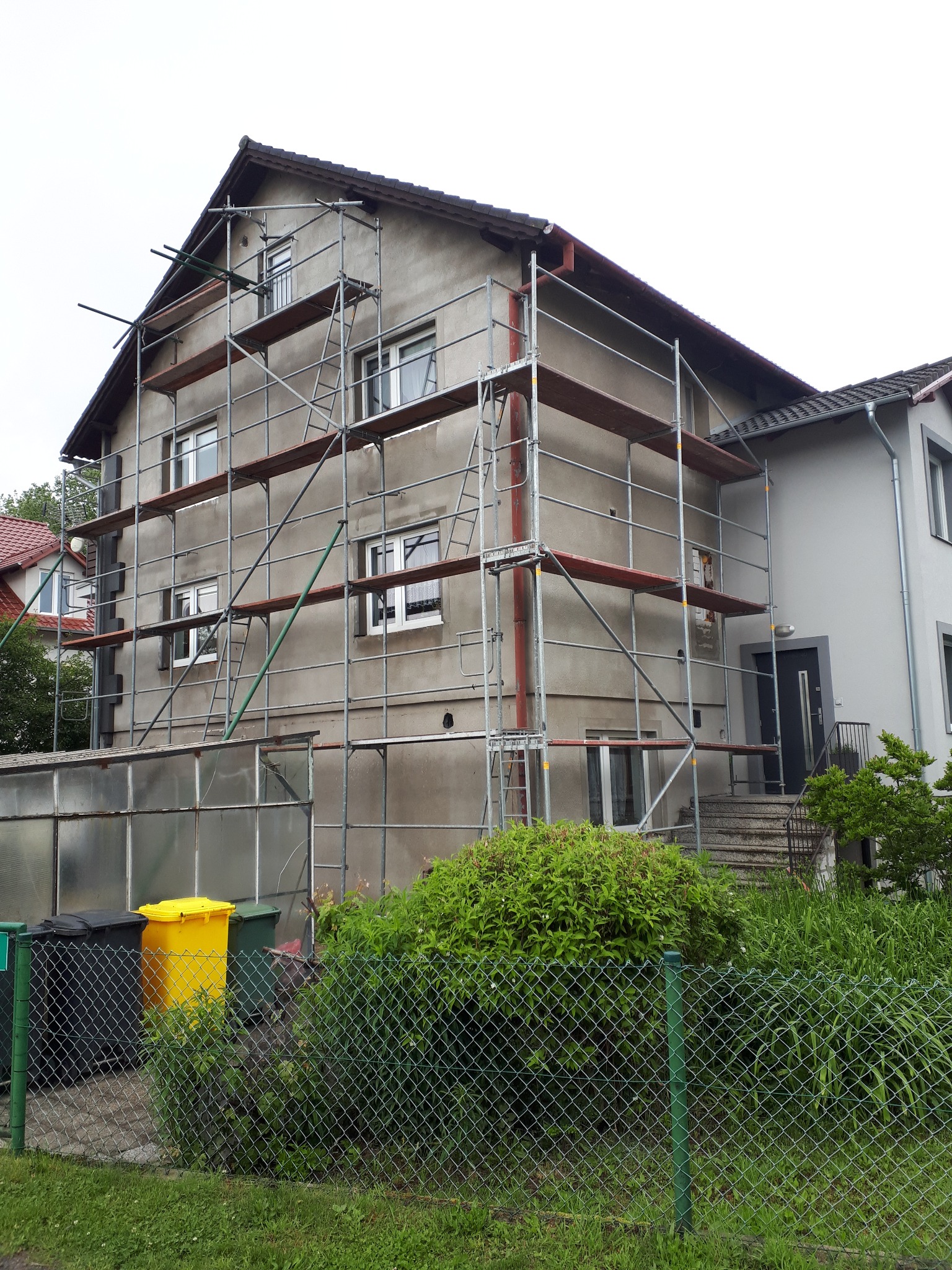 Dom w trakcie renowacji elewacji, otoczony rusztowaniem. Widoczne okna i fragment dachu. W tle zieleń i sąsiedni budynek.