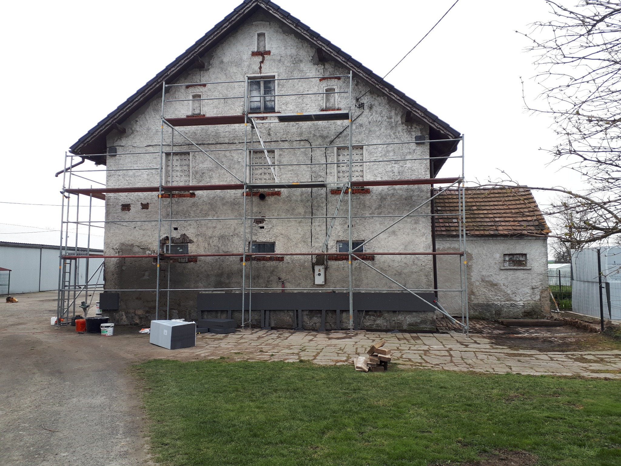 Stary dom z szarą elewacją w trakcie ocieplania, otoczony rusztowaniem. Część ściany pomalowana na szaro. Na trawie leżą resztki cegieł. Widoczne materiały budowlane.