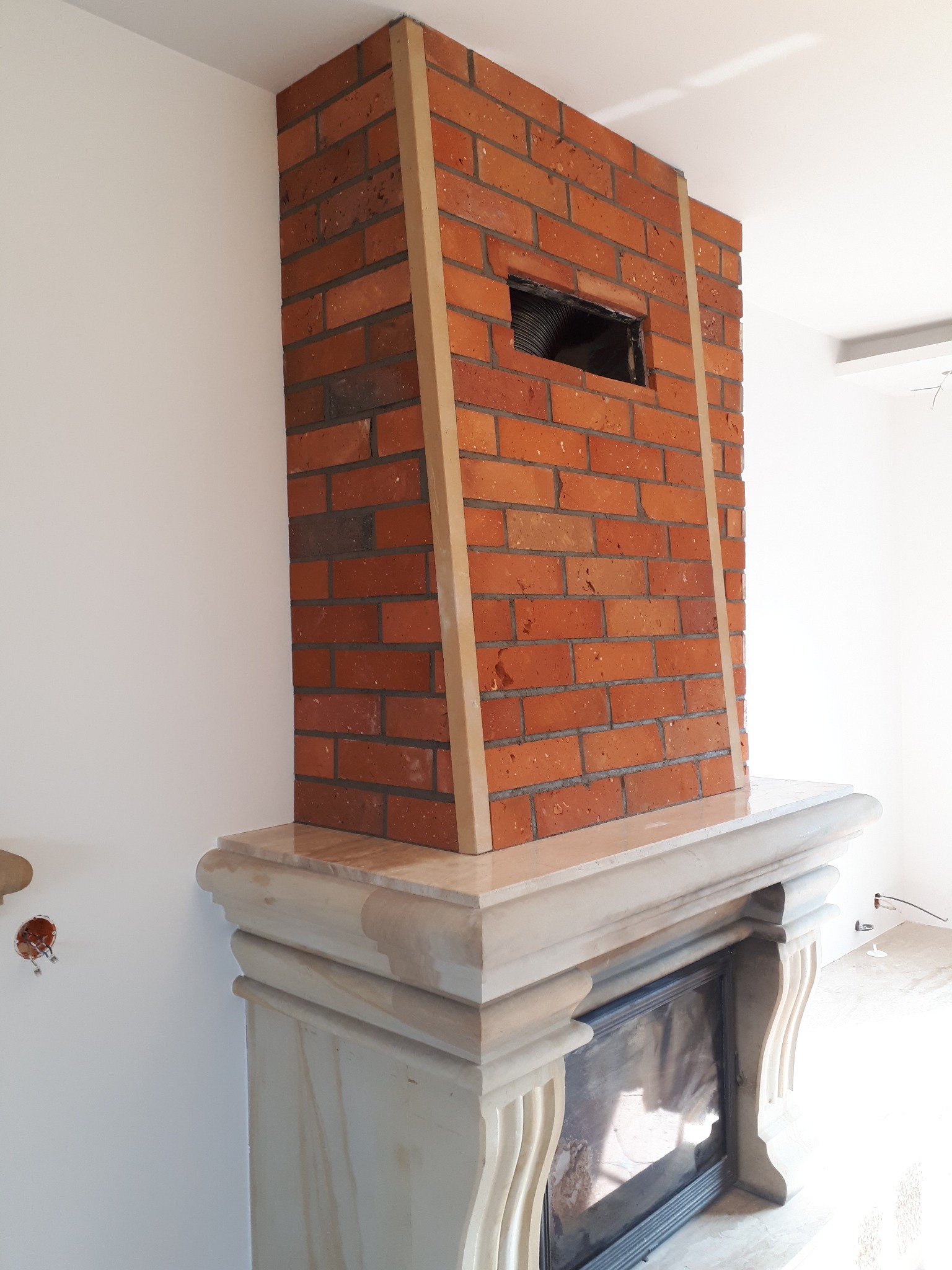 Murowany kominek z czerwonej cegły, obudowany jasnym kamieniem, z widocznym otworem wentylacyjnym i drewnianymi listwami wykończeniowymi, na tle białej ściany.