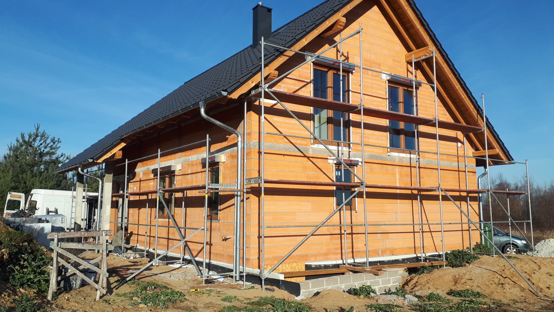 Dom w trakcie budowy: murowana konstrukcja z cegły, rusztowania, okna wstawione. Widoczne detale dachu i rynny. Elementy krajobrazu: drzewa, krzewy, błękitne niebo.