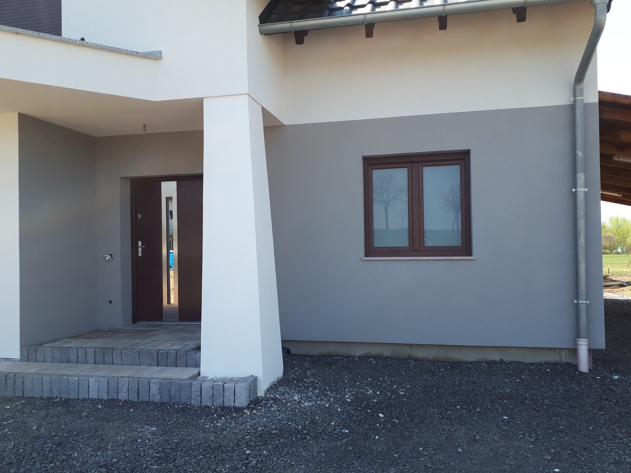 Dwukolorowa elewacja domu z brązowymi oknami i drzwiami wejściowymi. Widoczne schody z kostki brukowej i szara elewacja w dolnej części budynku.