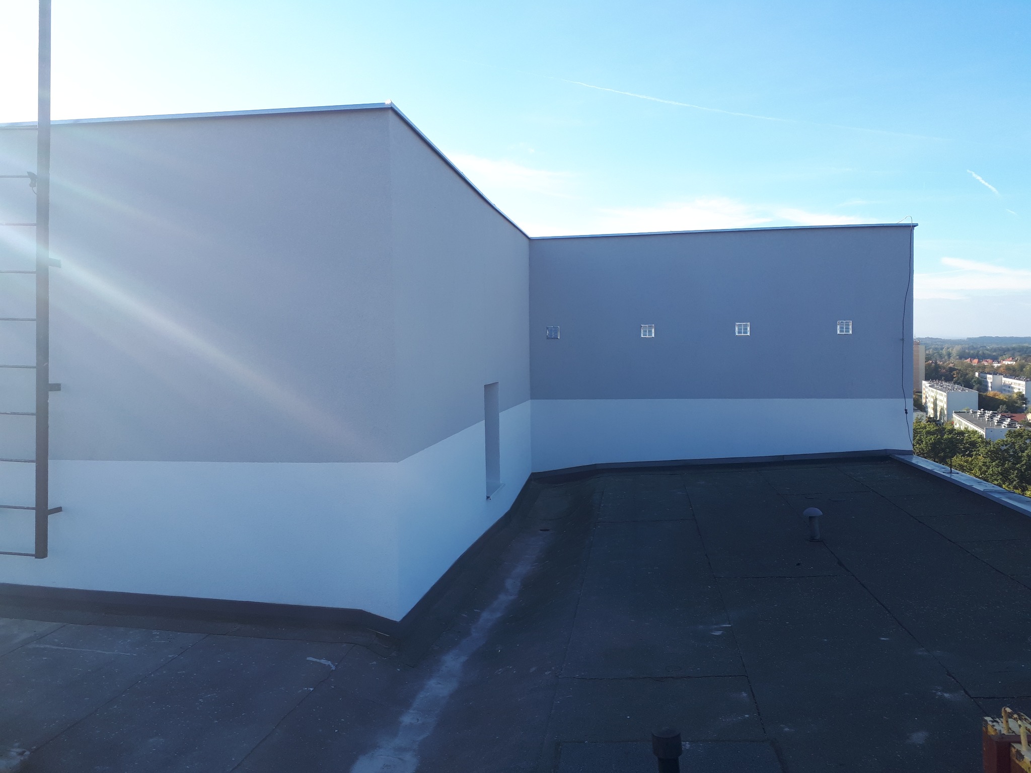 Elewacja budynku po malowaniu z drabinką i małymi okienkami wentylacyjnymi, widok z dachu w słoneczny dzień. Jasnoszara góra i biały dół elewacji.
