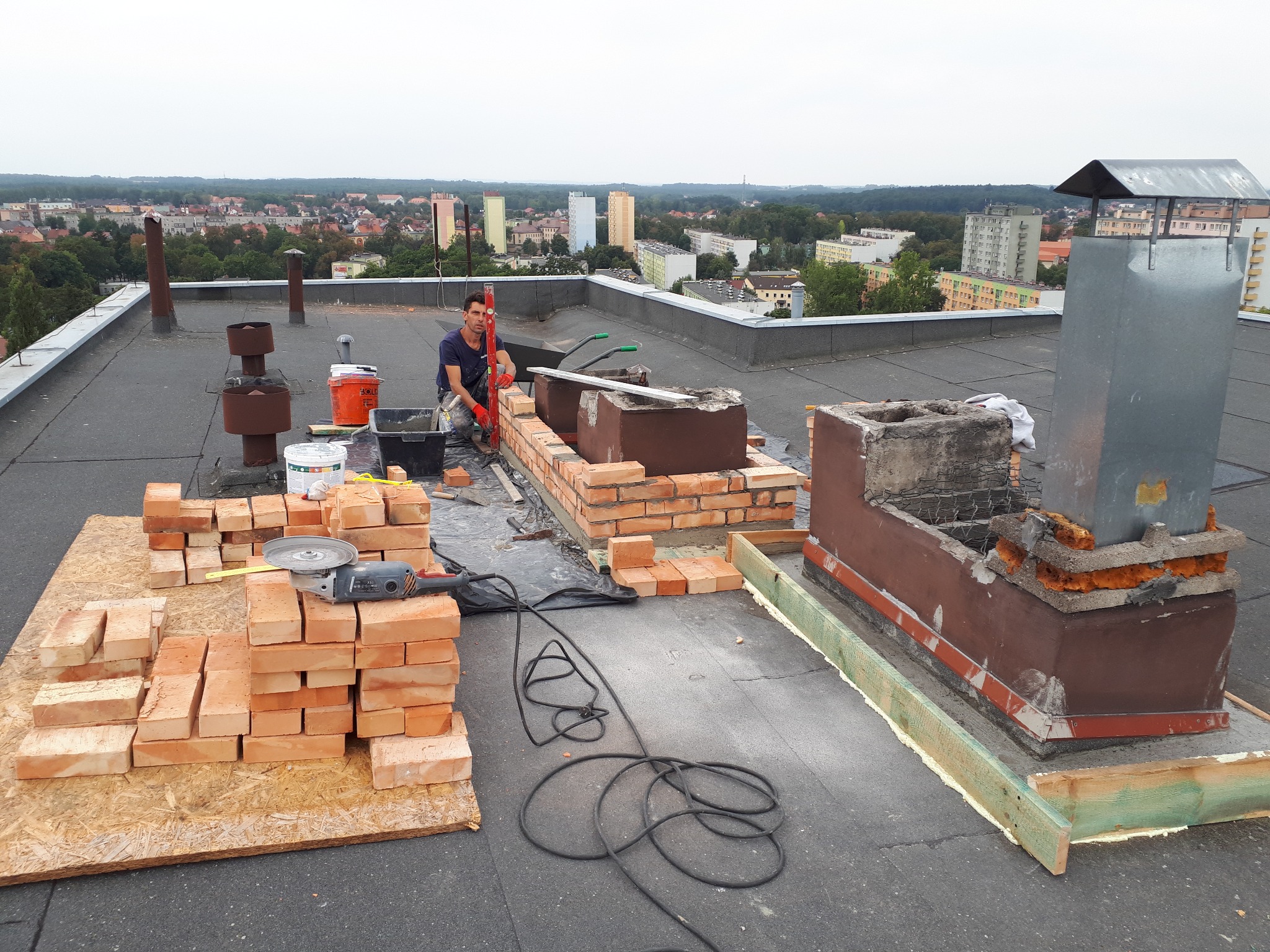 Murarz w trakcie budowy komina na dachu budynku. Widoczne cegły, narzędzia i panorama miasta w tle. Prace murarskie z użyciem poziomicy i zaprawy.