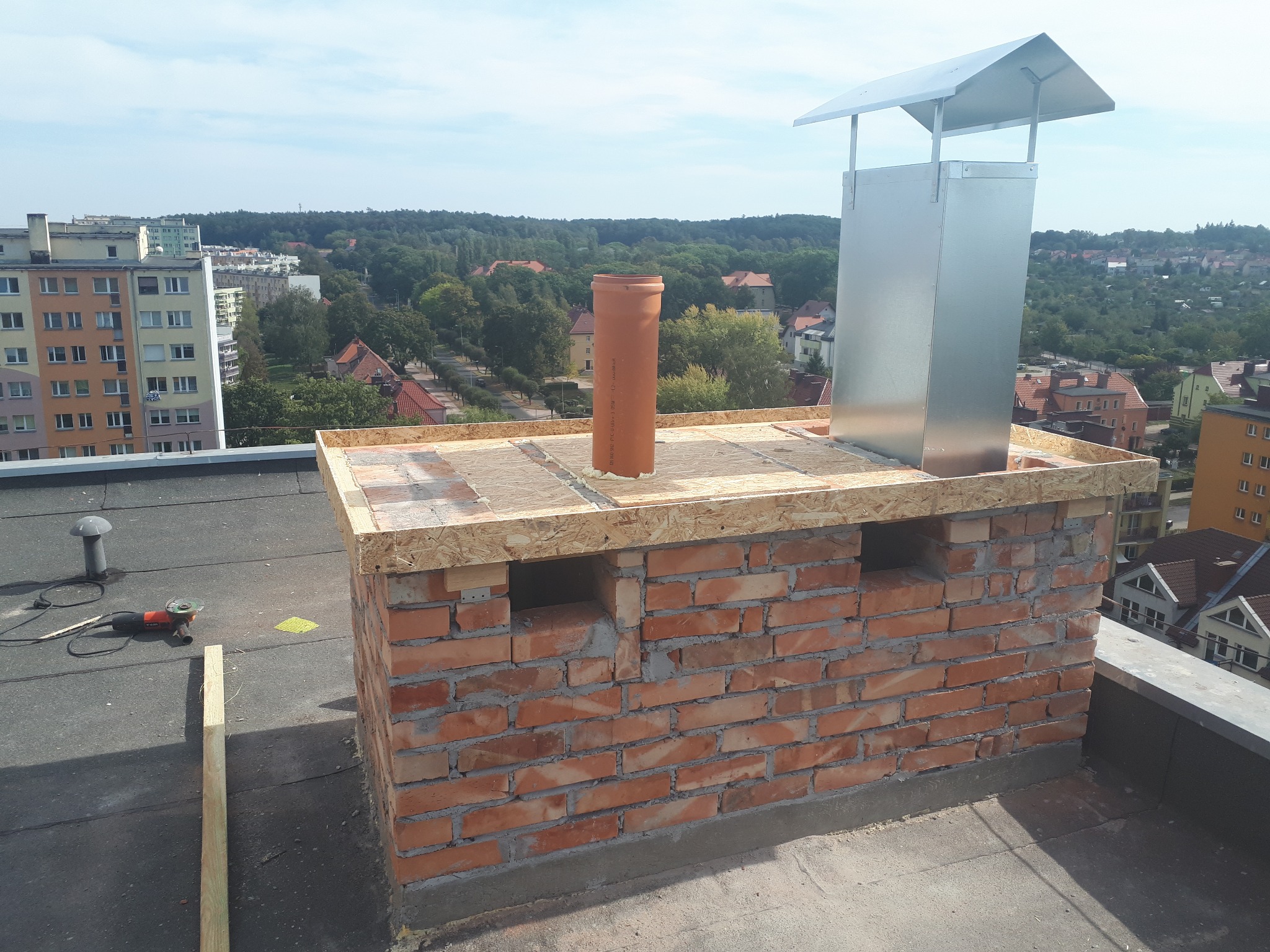 Wznoszony z cegły komin z metalowym wkładem i daszkiem na dachu budynku, widok na miasto w tle. Na dachu leży szlifierka kątowa.