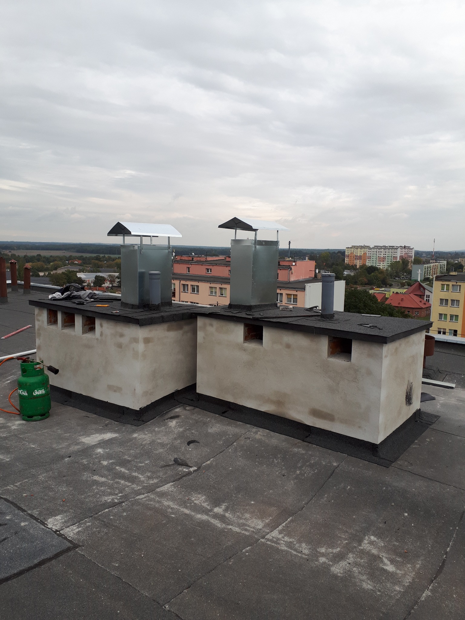 Dwa kominy na dachu budynku z nowymi, metalowymi zakończeniami. Widoczna butla gazowa i narzędzia. W tle zabudowa miejska i pochmurne niebo.