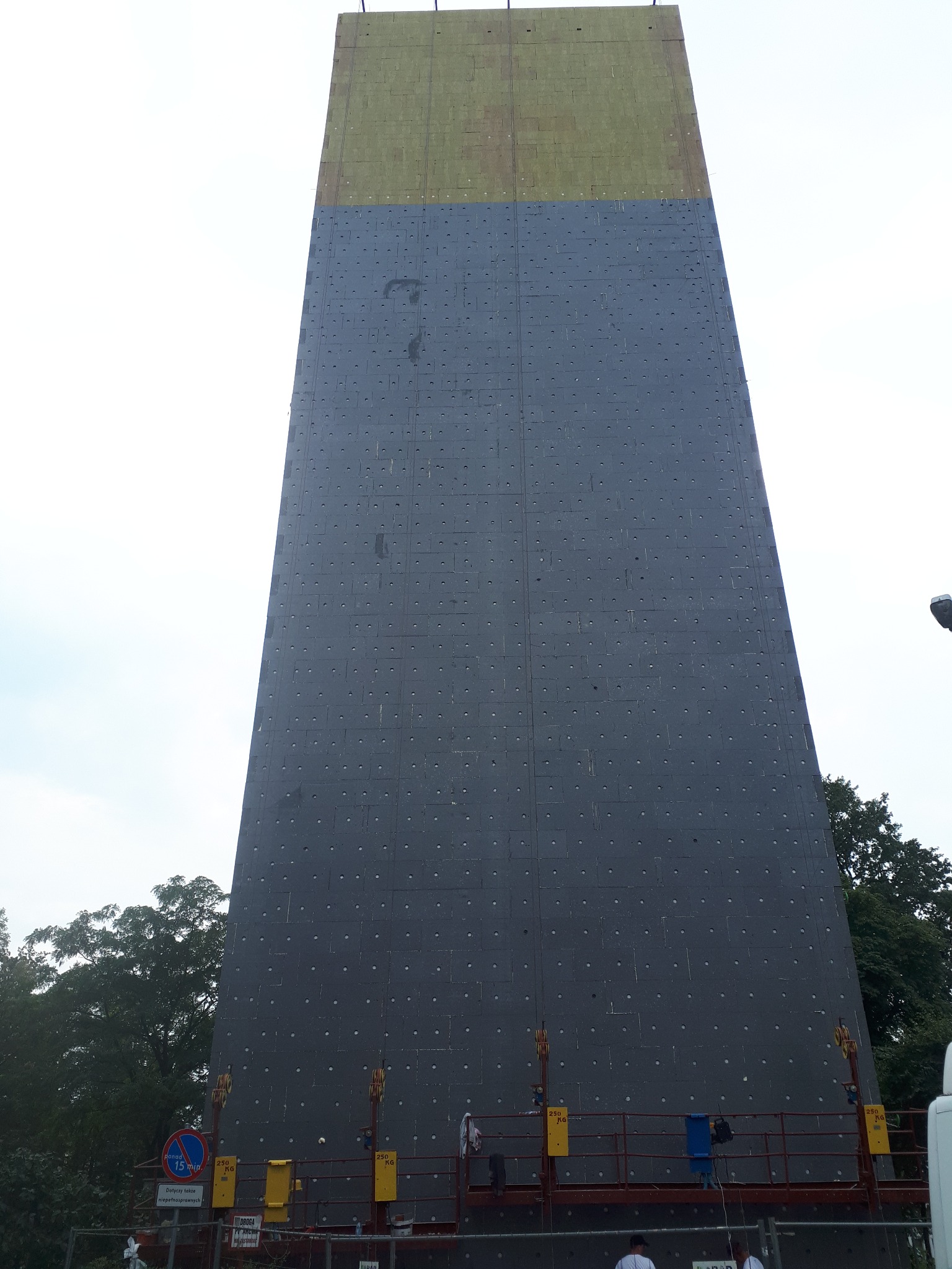 Wysoka ściana budynku w trakcie ocieplania, z widoczną szarą warstwą izolacyjną i górną częścią pokrytą żółtym materiałem izolacyjnym. Prace na wysokości.