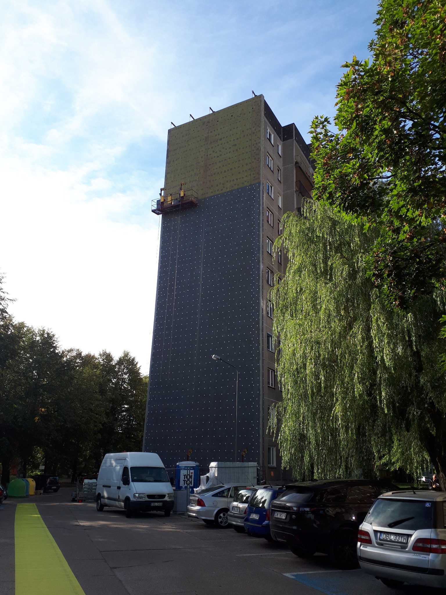 Ocieplanie elewacji budynku wielorodzinnego z użyciem rusztowania wiszącego; widoczne płyty izolacyjne i siatka z otworami. Na dole parkujące samochody i drzewo.