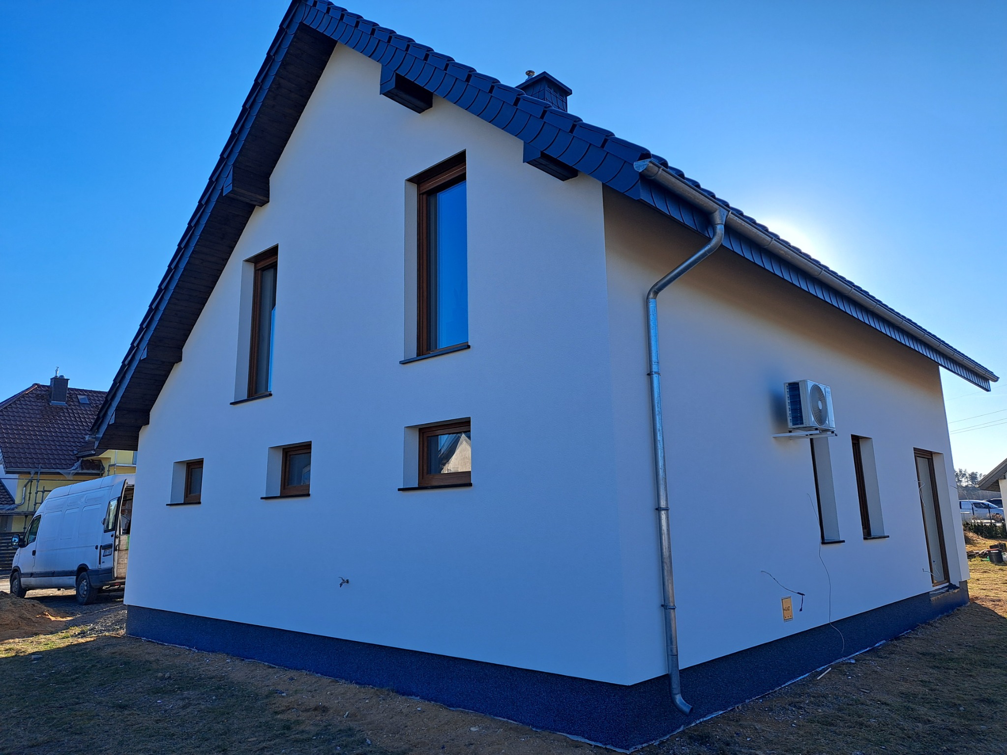 Biały dom jednorodzinny z ciemną dachówką i drewnianymi oknami, widok z boku, na tle błękitnego nieba i zaparkowanego białego busa. Widoczny klimatyzator na elewacji.