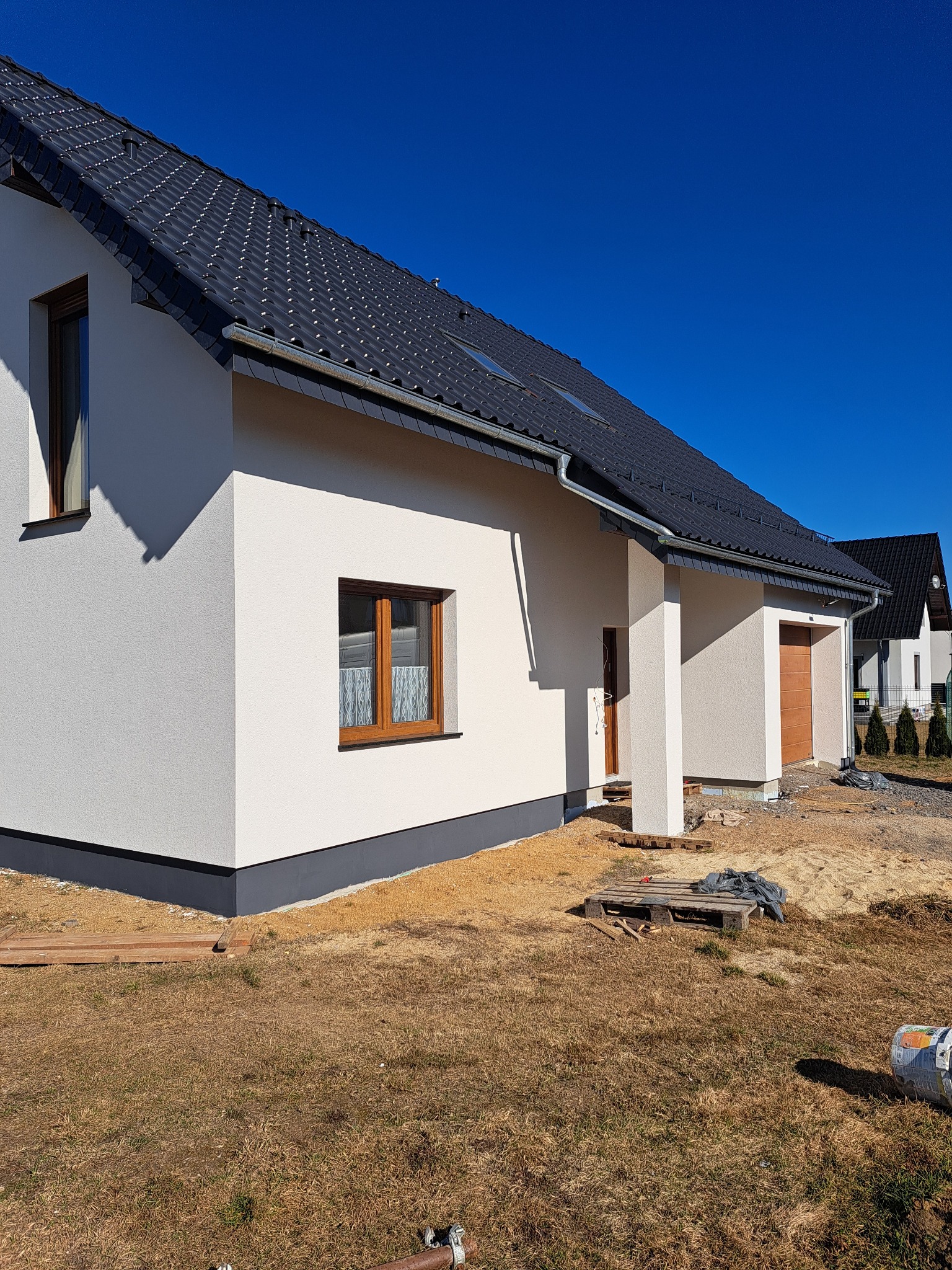 Nowoczesny dom jednorodzinny z białą elewacją i grafitowym dachem w słoneczny dzień. Widoczne okna z drewnianą stolarką i fragment niezagospodarowanego terenu.