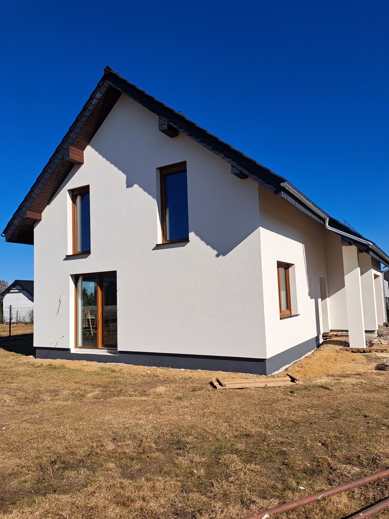 Biały dom jednorodzinny z brązowymi oknami i ciemnym dachem na tle błękitnego nieba. Widoczne elementy wykończeniowe i fragment terenu.