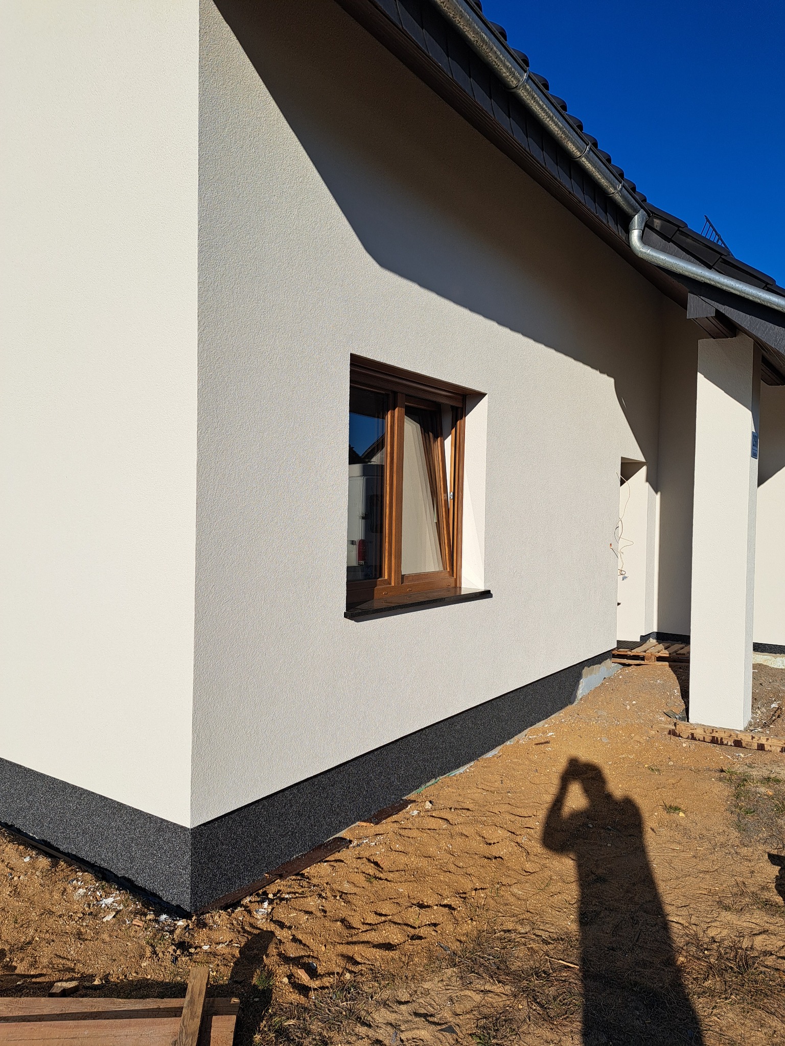 Jasna elewacja domu jednorodzinnego z brązowymi oknami i ciemną podmurówką. Widoczny fragment dachu i cień osoby robiącej zdjęcie. Słoneczny dzień.